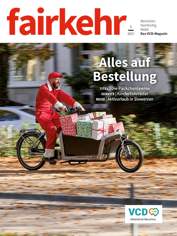 Auf dem Cover der fairkehr 5/2021 zum Thema Citylogistik fährt ein Mann im roten Overall ein Lastenfahrrad voller Weihnachtsgeschenke.